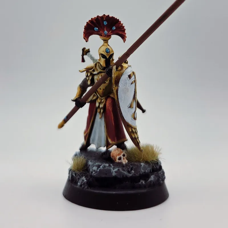 Un guerrier à la lance, en armure dorée et cape rouge, sur un socle rocheux parsemé de crânes.
